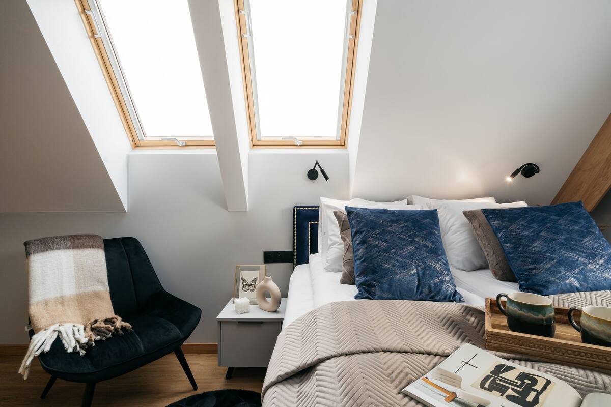 A modern bedroom with a skylight, a bed with blue and white bedding, a dark armchair, and a bedside table with a vase and picture frame.