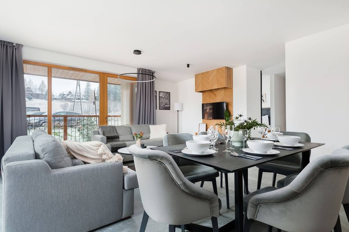 A modern living room with a dining area, grey sofas, a dark dining table, and large windows with curtains.