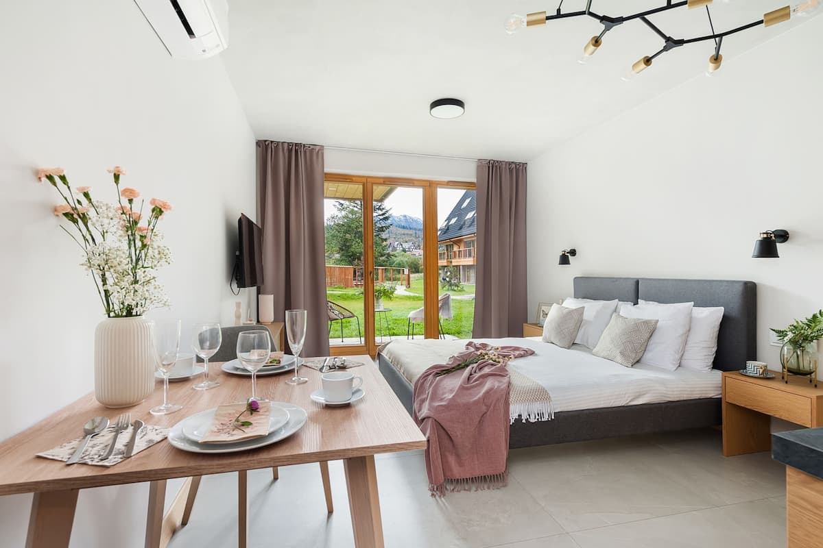 A modern bedroom with a large bed, a dining table set for two, and a view of a garden and mountains.