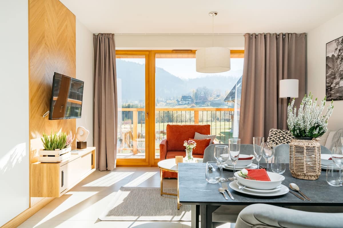 A modern living room with a dining area, large windows, and a view of mountains.