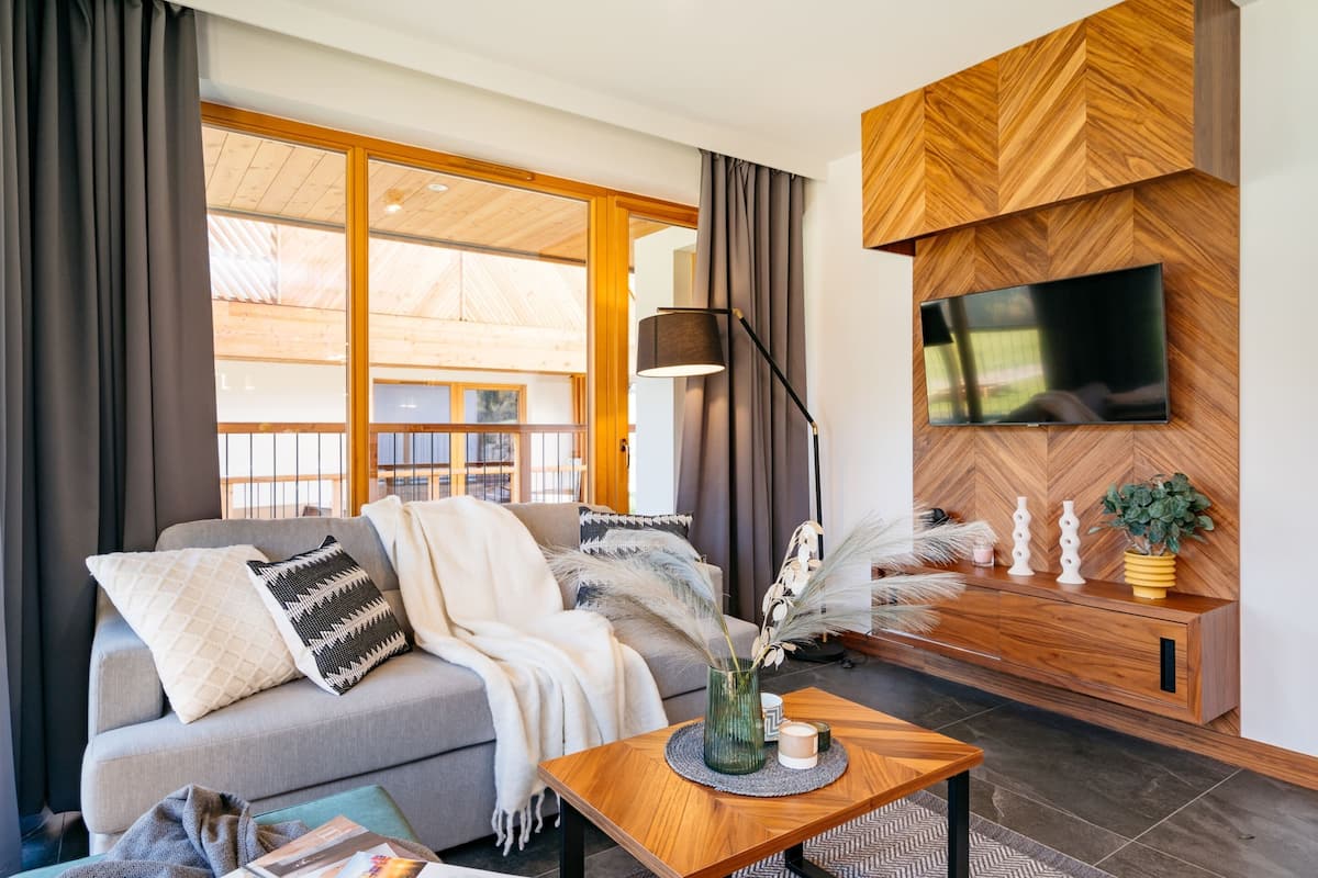 A modern living room with a sofa, a coffee table, a TV mounted on a wooden wall, and a large window with curtains.