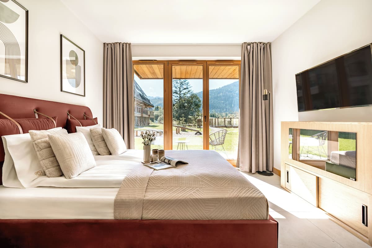 A modern bedroom with a large bed, a flat-screen TV, and a view of a mountainous landscape through large windows.