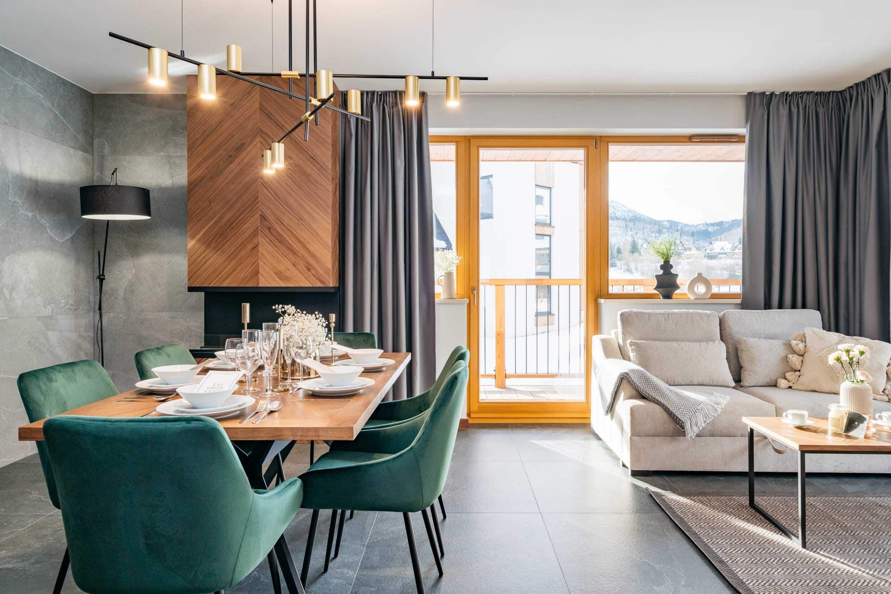 Un comedor moderno con una mesa de madera dispuesta para cuatro personas, sillas tapizadas de color verde y una lámpara colgante contemporánea.