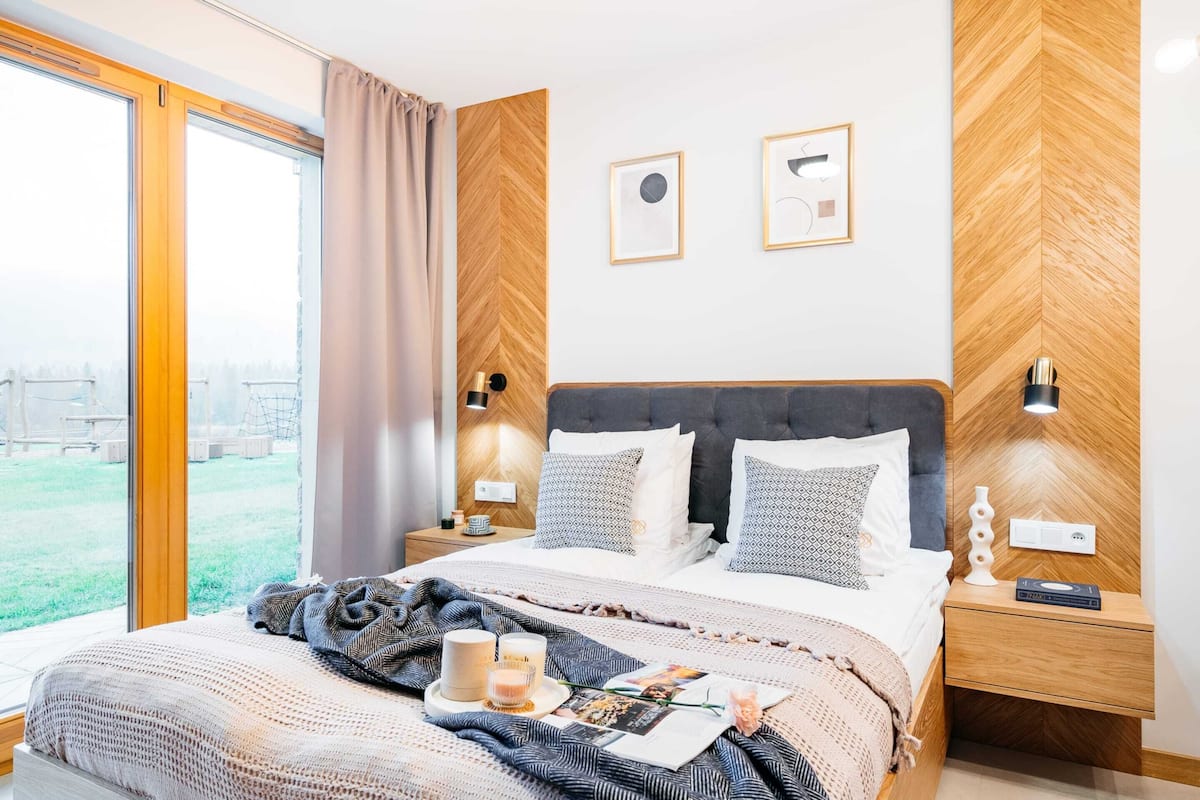 A modern bedroom with a large bed, wooden headboard, two bedside tables with lamps, and a view of a grassy area through large windows.