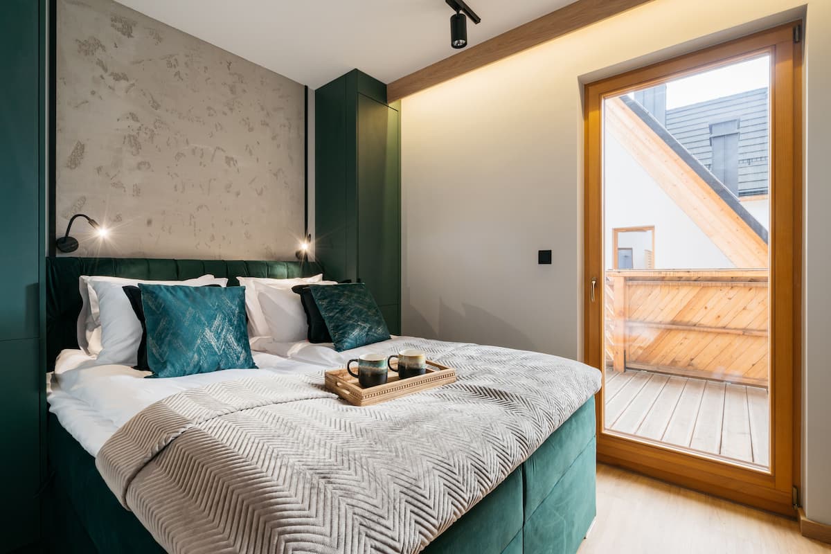 A bedroom with a bed, pillows, a tray with cups, a wooden ceiling, and a view of a wooden deck through a glass door.