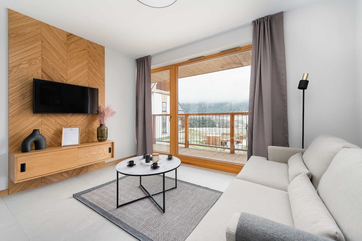 A modern living room with a flat-screen TV, a sofa, a coffee table, and a large window with a view of a balcony.