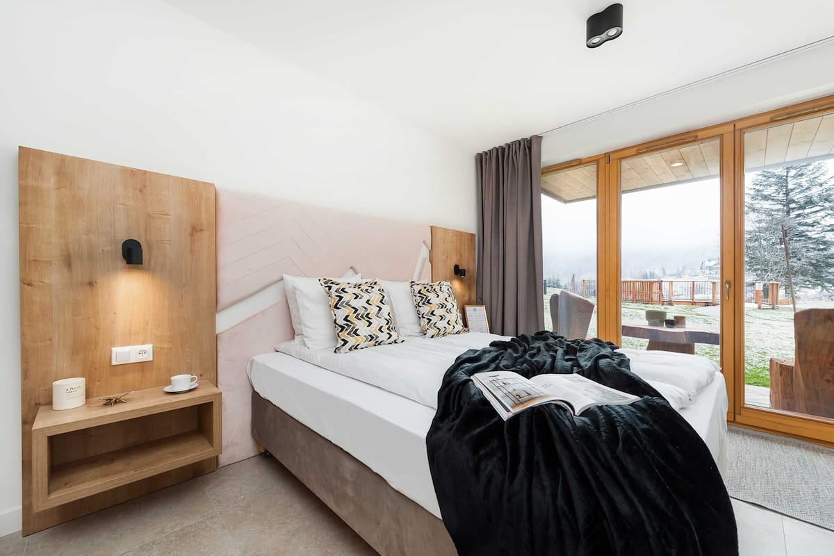 A modern bedroom with a wooden headboard, a bedside table with a cup and a book, and a large window with a view of a snowy landscape.