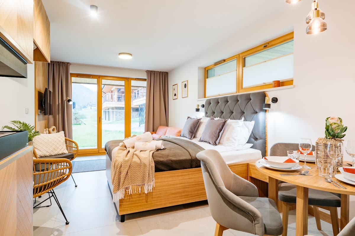 A modern hotel room with a bed, a dining table with chairs, a TV, and a view of the outdoors.