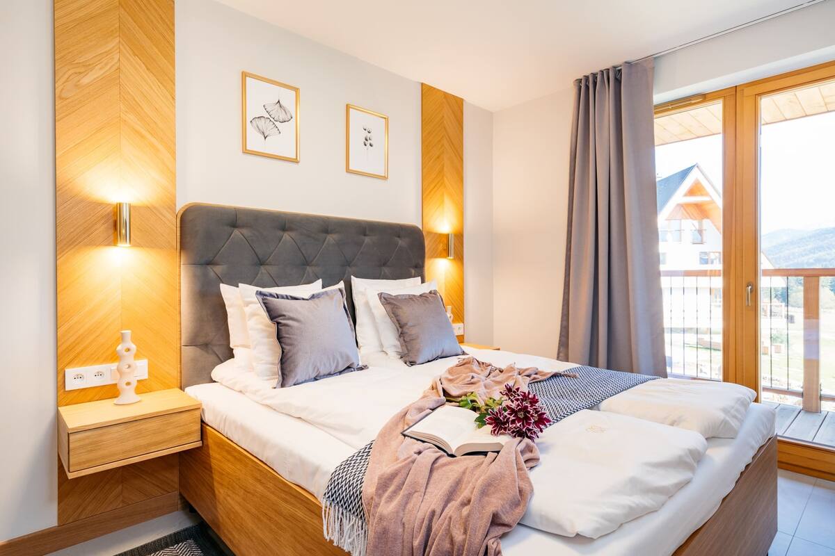 A modern bedroom with a large bed, wooden headboard, bedside lamps, and a view of a building through a large window.