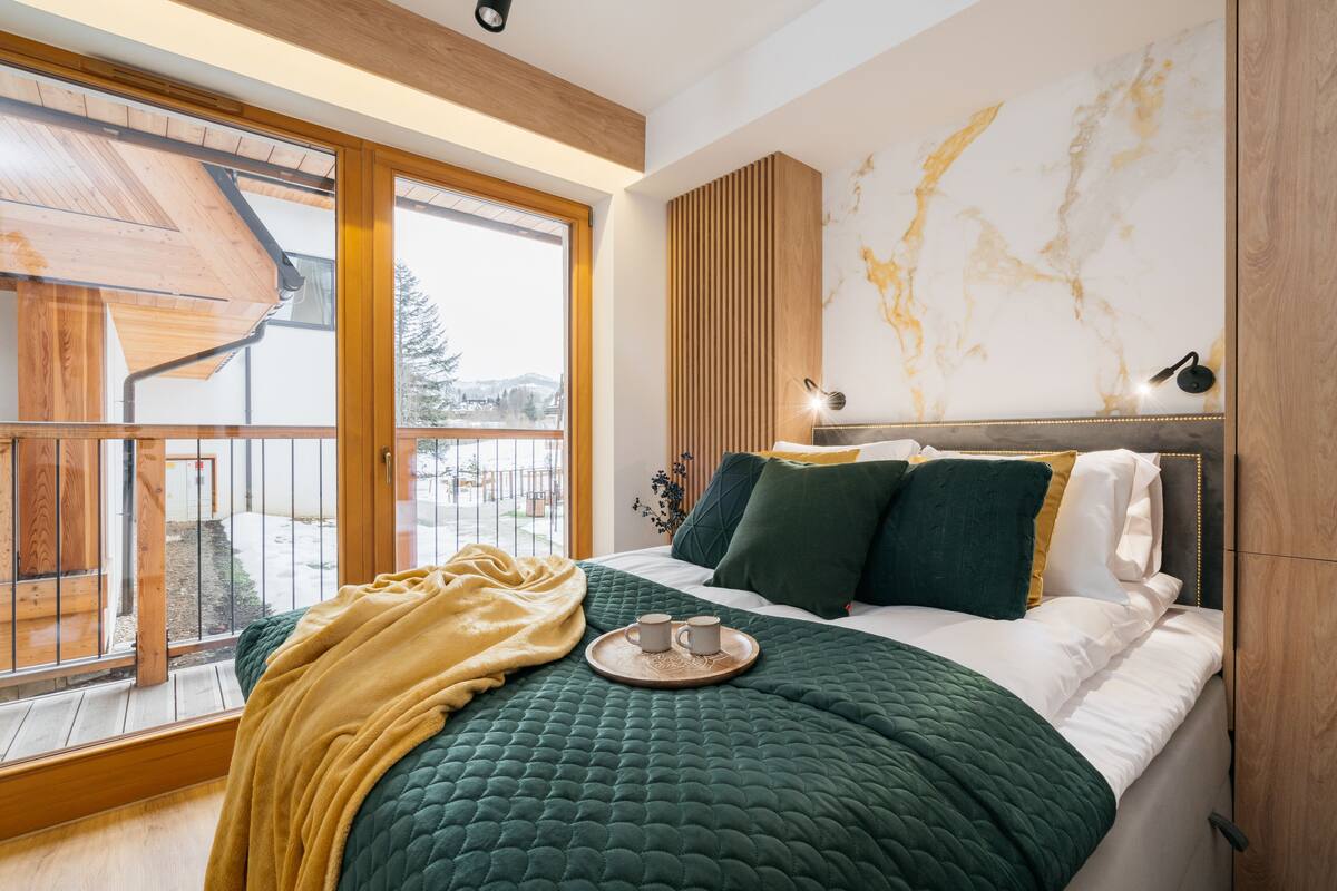 A modern bedroom with a large bed, a wooden headboard, a marble wall, and a view through a sliding glass door.