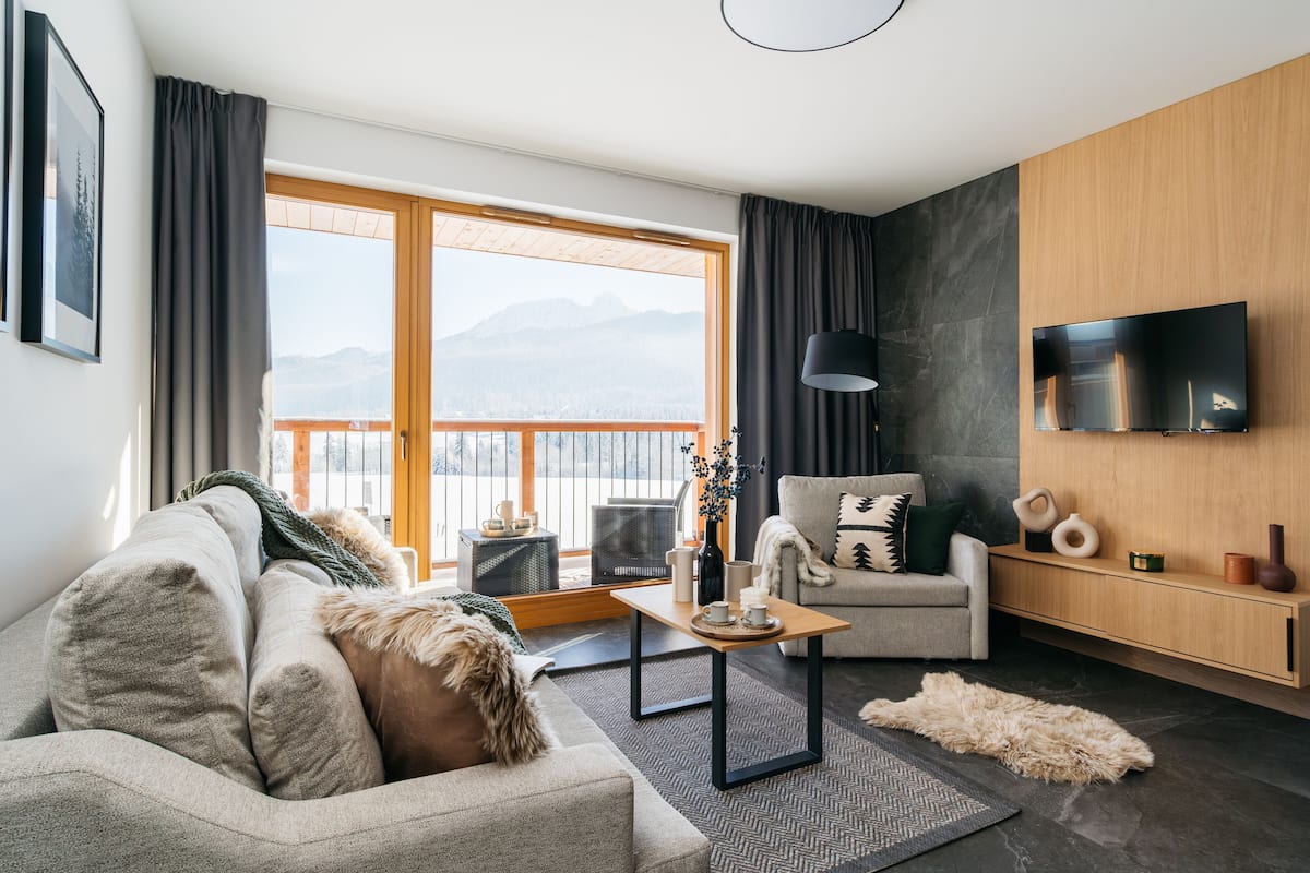 A modern living room with a sofa, a coffee table, a chair, a TV mounted on the wall, and a view of mountains through large windows.