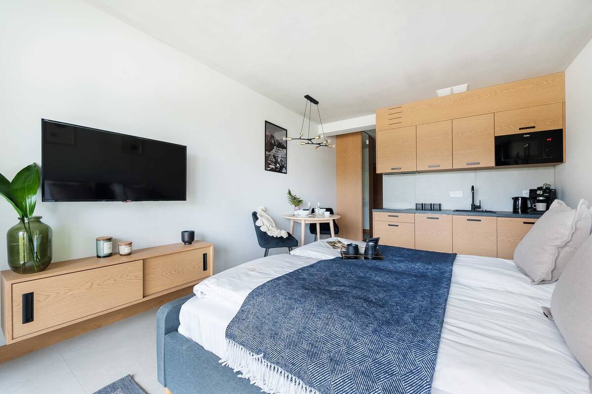 A modern hotel room with a large bed, a TV mounted on the wall, a small kitchen area, and a seating area with a table and chair.