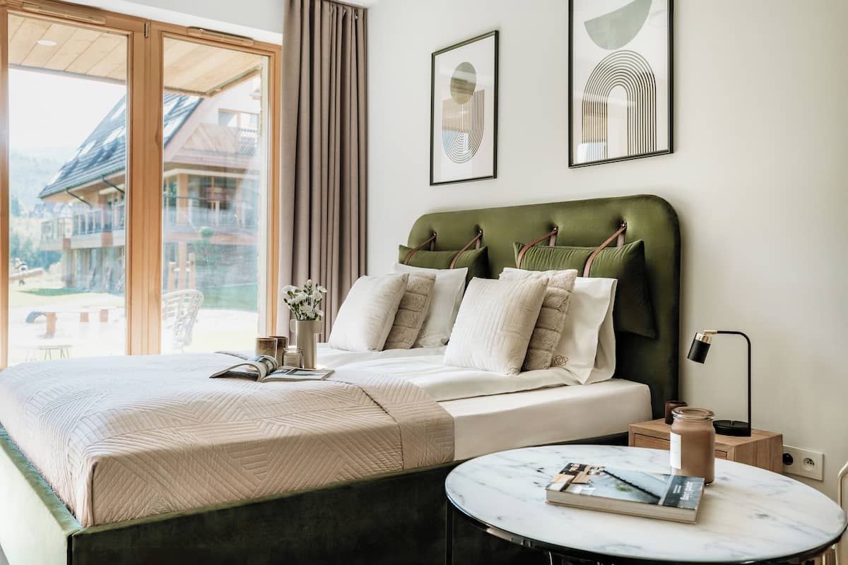 A neatly made bed with a green headboard, a bedside table with a book and a candle, and a window with curtains.