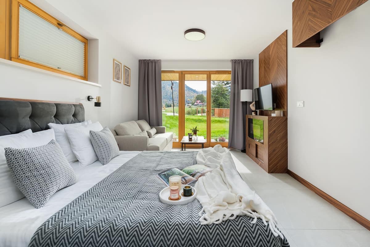 A modern bedroom with a large bed, a sofa, a TV, and a view of a green landscape.