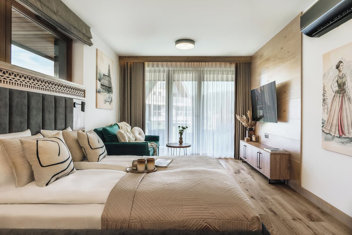 A modern hotel room with a large bed, a green sofa, a small table with a vase, and a mounted TV.