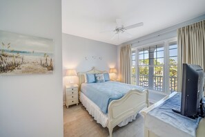 A bedroom with a bed, bedside lamps, a ceiling fan, a TV on a desk, and a large window with curtains.