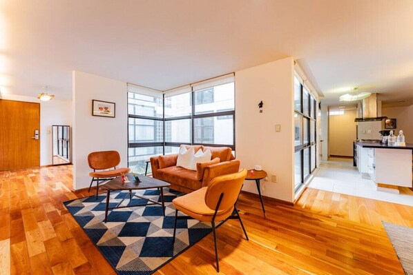 Apartamento moderno com sala de estar, incluindo sofá, cadeiras e mesa de centro. Cozinha visível através de uma porta aberta.