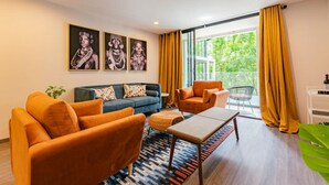 Een moderne woonkamer met een bank, fauteuils en een salontafel. Er hangen drie ingekaderde foto's aan de muur, een plant en een balkon met gele gordijnen.