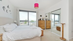 A bedroom with a large bed, a dresser, a mirror, and a view of the city through large windows. - Putney Rooftops (London)