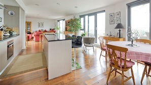 A modern kitchen with wooden flooring, a dining area with a checkered tablecloth, and a view of the city through large windows. - Putney Rooftops (London)