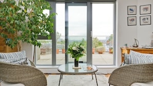 A modern living room with wicker furniture, a glass table with a vase of flowers, and a large sliding glass door leading to a balcony with potted plants. - Putney Rooftops (London)