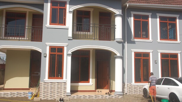 A multi-story house with a balcony, a car parked in front, and a person standing near the entrance. - MiGo House (Arusha)