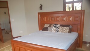 A wooden bed with a white mattress and patterned pillows in a room with a tiled floor and a door in the background. - MiGo House (Arusha)