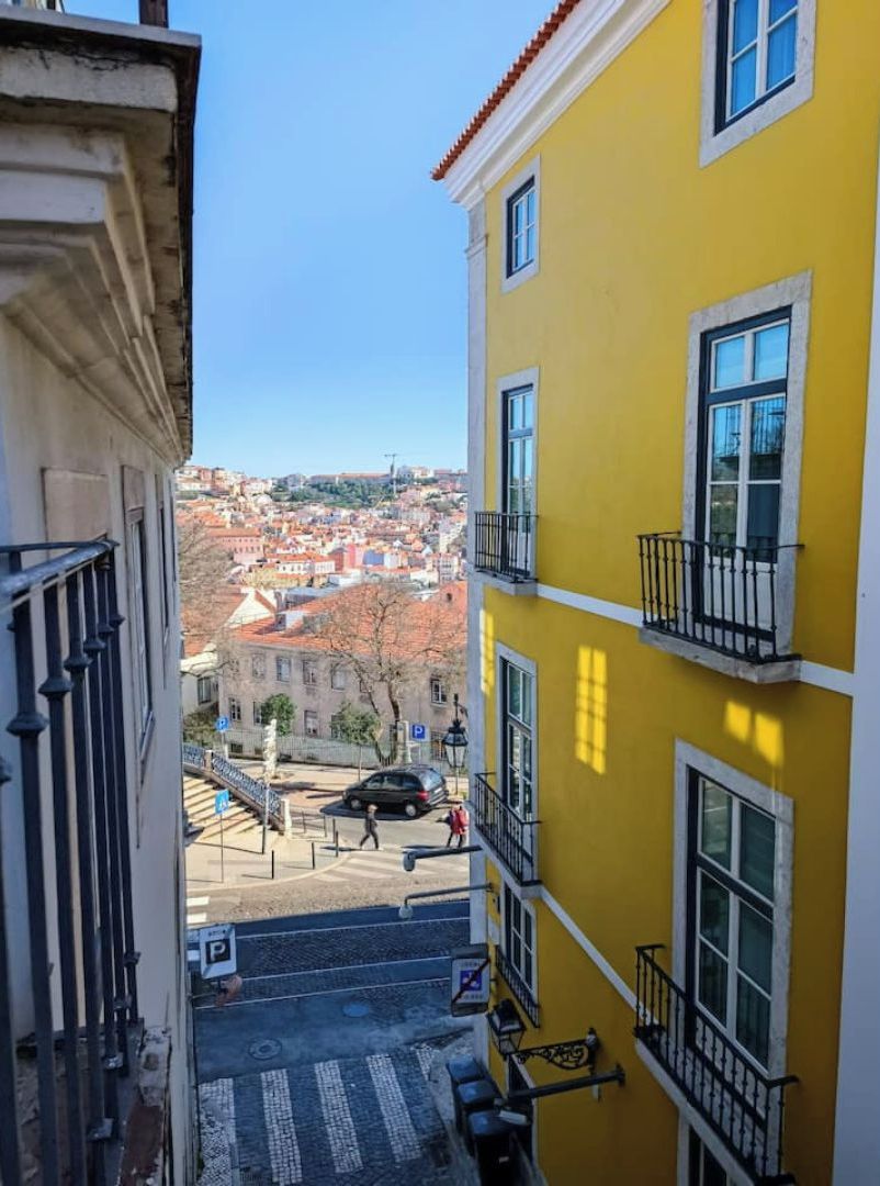 A narrow street with a yellow building on the right and a white building on the left. There are steps leading up to a balcony with a railing.