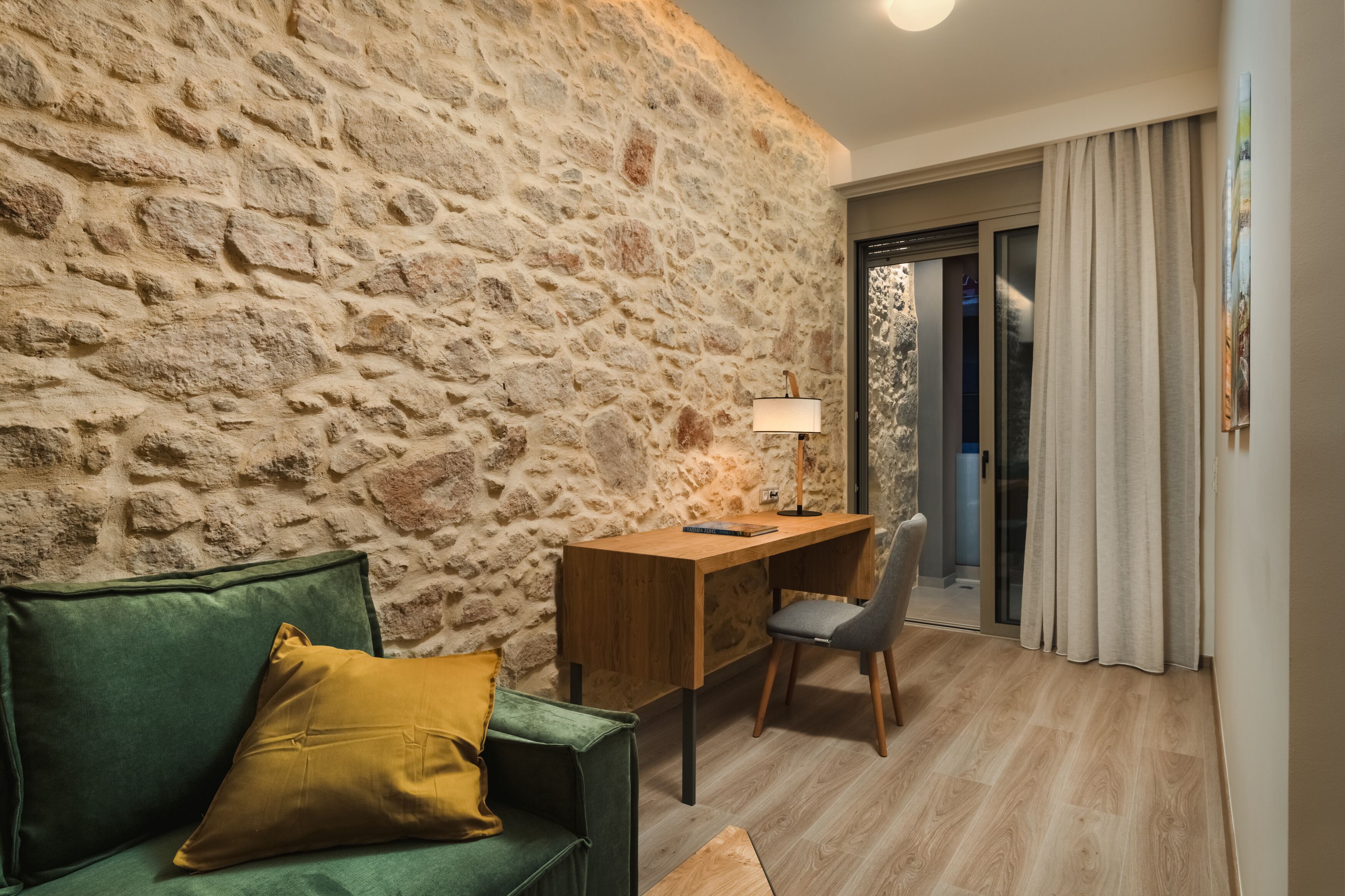A room with a stone wall, a wooden desk with a chair, a green sofa, and a window with curtains.