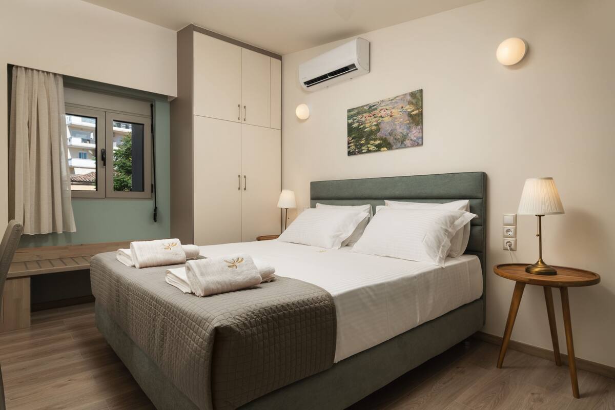 A neatly made bed with white linens and a green headboard, a bedside table with a lamp, and a window with curtains.