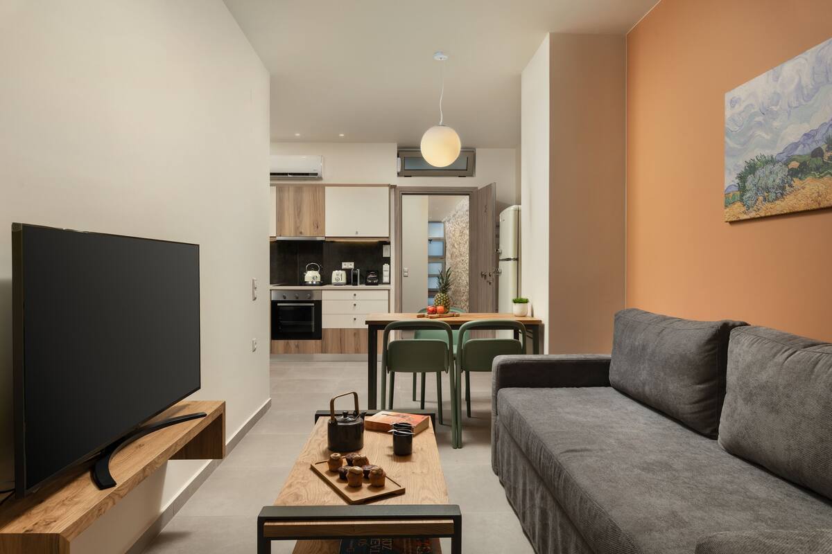 A modern living room with a flat-screen TV, a wooden coffee table, a grey sofa, and a dining area with a table and chairs.