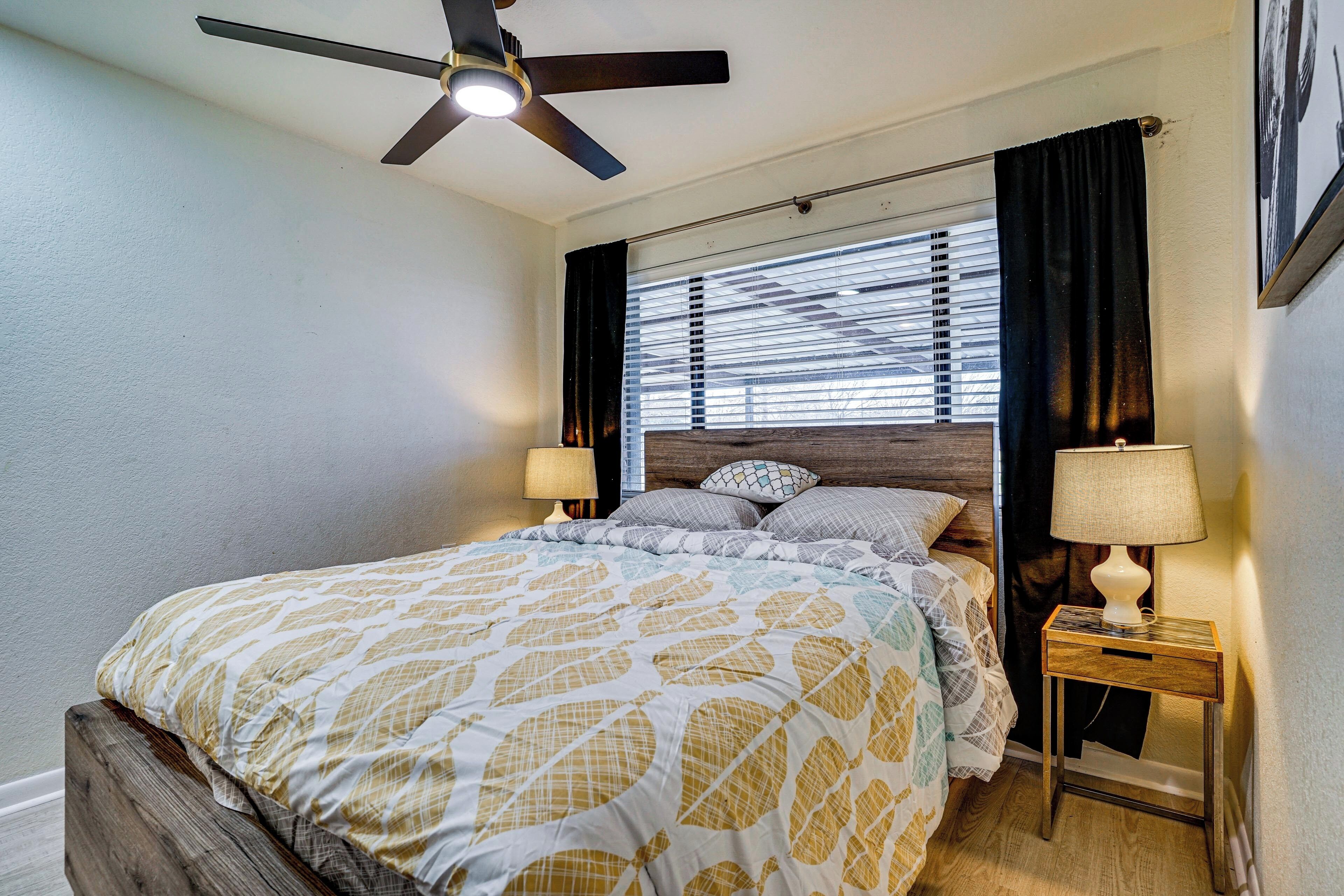 A bedroom with a bed, a nightstand, a lamp, a ceiling fan, and a window with blinds.