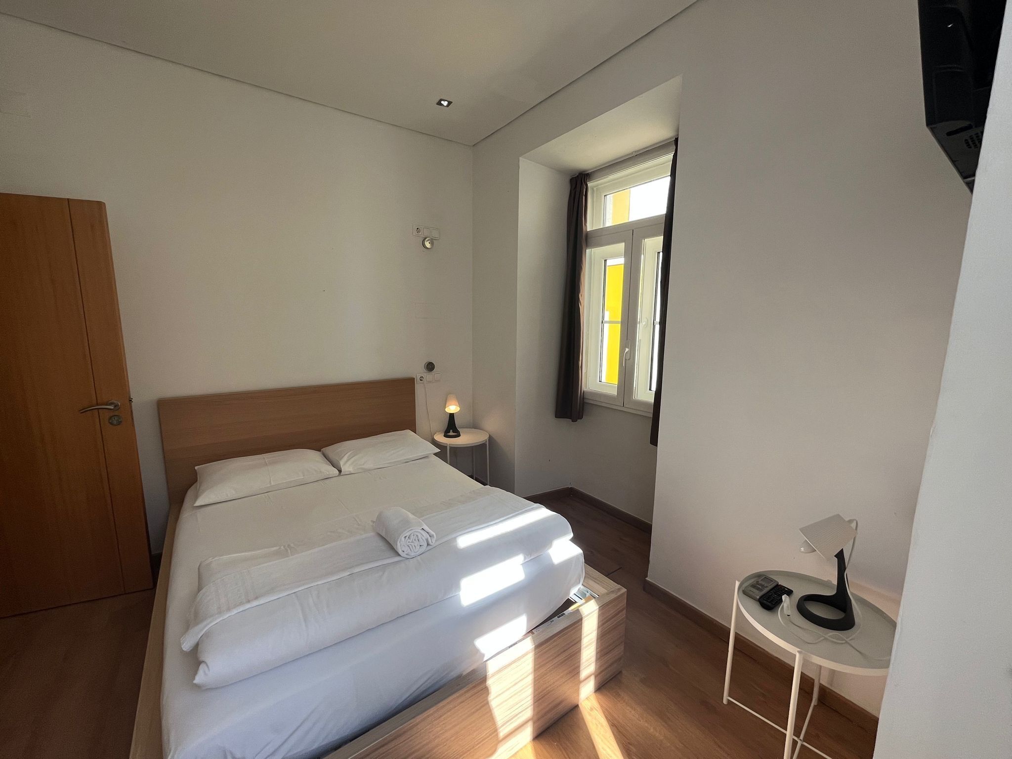 A neatly made bed with a white duvet and pillows, a bedside table with a lamp, and a small table with a phone and charger.