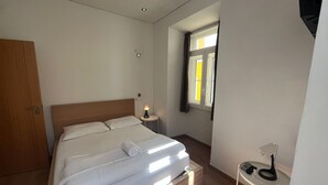 A neatly made bed with a white duvet and pillows, a bedside table with a lamp, and a small table with a phone and charger. - Bairro Alto - 3 bedroom apartment with 2,5 bathrooms (Lisboa)