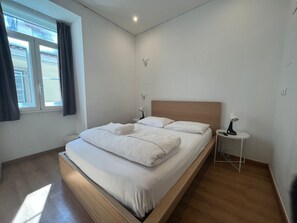 Une chambre à coucher moderne avec un lit en bois, une literie blanche, une table de chevet avec une lampe et une fenêtre avec des rideaux.