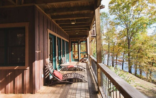 Relax on the covered porch with two lounge chairs and a small table, surrounded by a serene natural setting and a body of water.