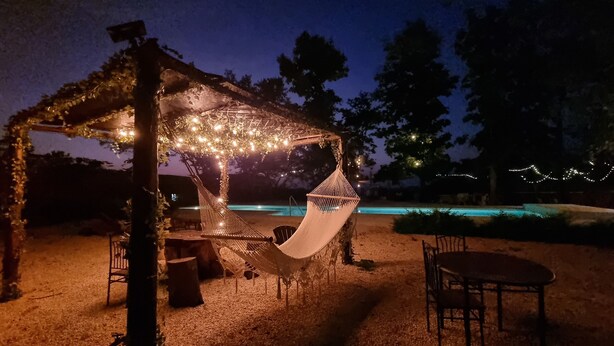 Un hamac sous une pergola illuminée de guirlandes lumineuses, une piscine et un espace salon extérieur.