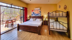 Une chambre avec un lit en bois, un ventilateur de plafond, un tableau, un lit superposé et une vue sur une forêt, visible à travers de grandes fenêtres.