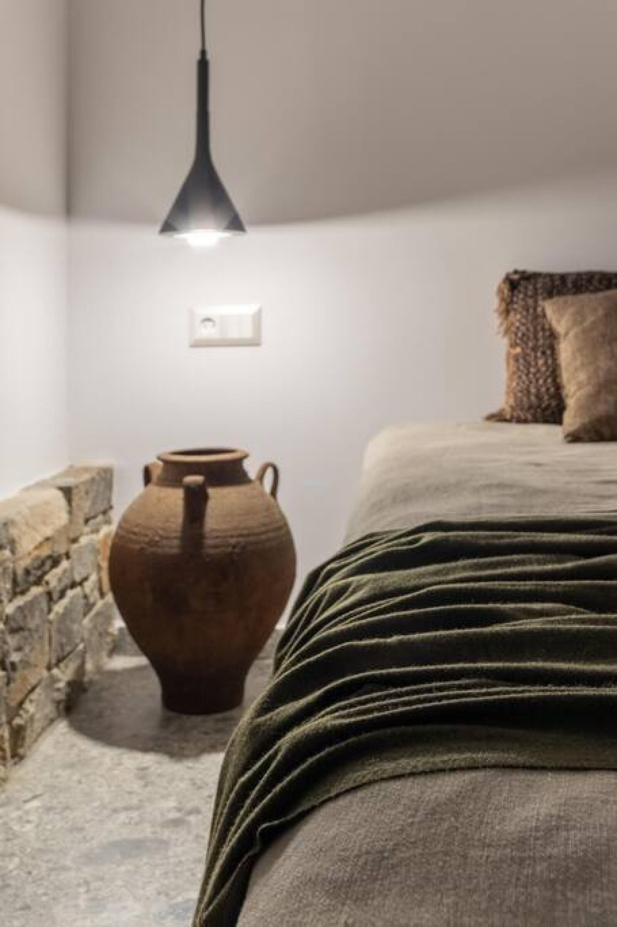Una camera da letto con un letto, una lampada a sospensione, un muro in pietra e un vaso di ceramica di grandi dimensioni.