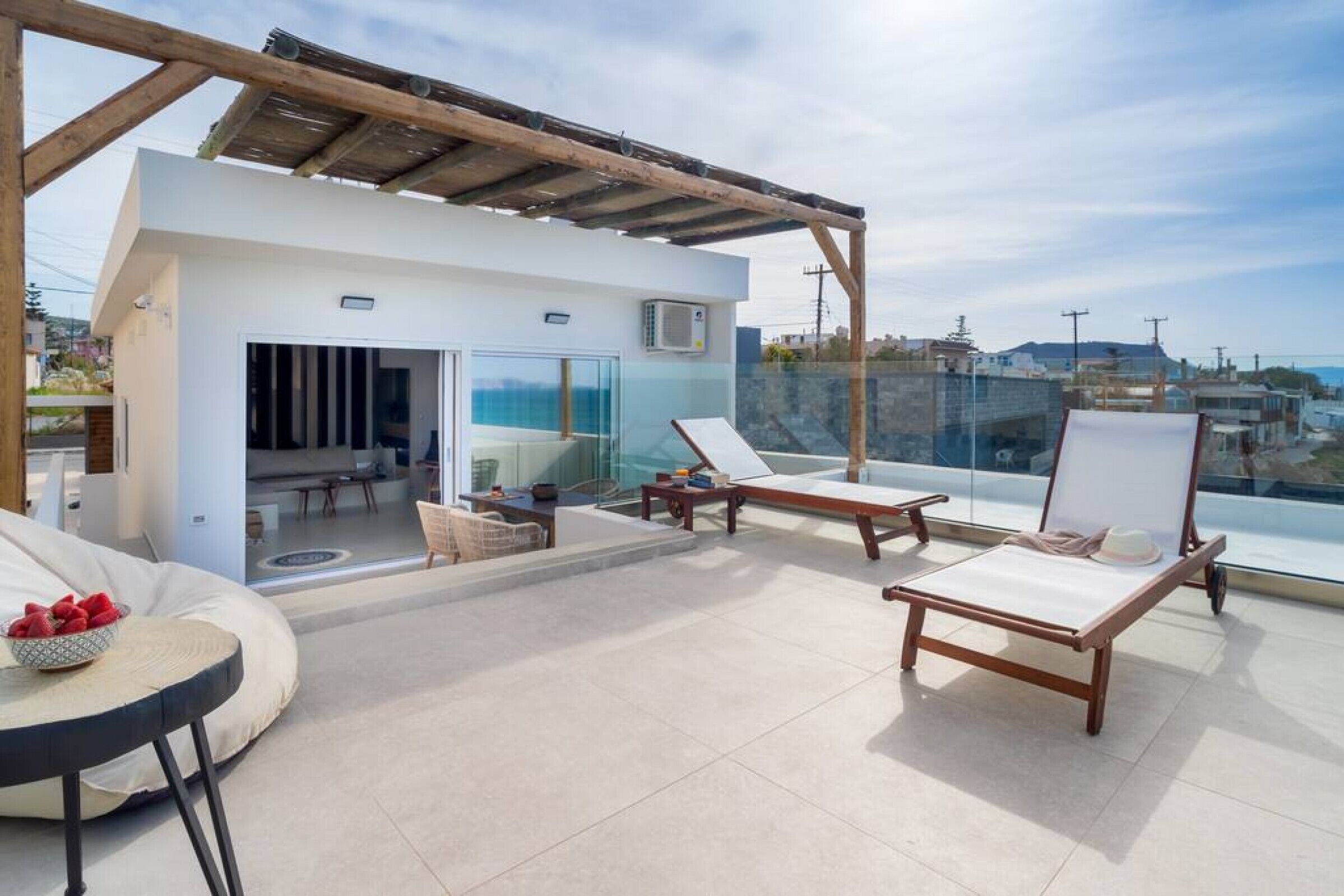 A rooftop terrace with a modern design, featuring a glass door leading to an indoor area, two lounge chairs, and a small table with a bowl of fruit.