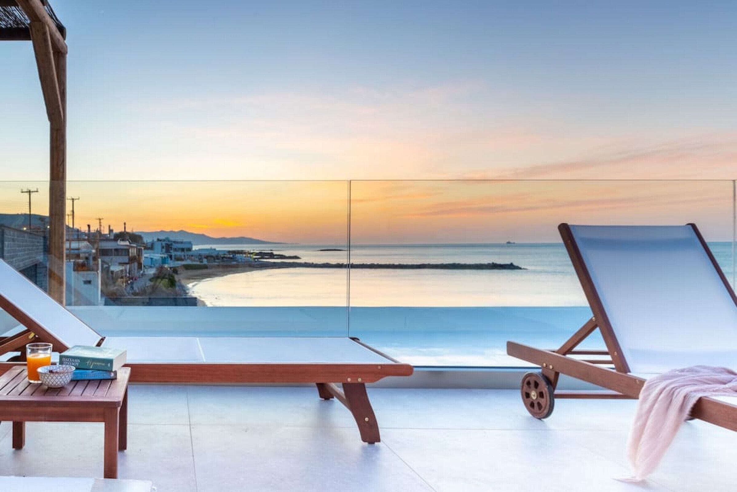 Un balcone con due poltrone, un tavolo con un bicchiere di succo d'arancia e un libro, e vista sul mare e sugli edifici in lontananza al tramonto.