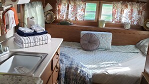 A compact RV interior with a kitchenette, a bed with a patterned bedspread, and a window with curtains.