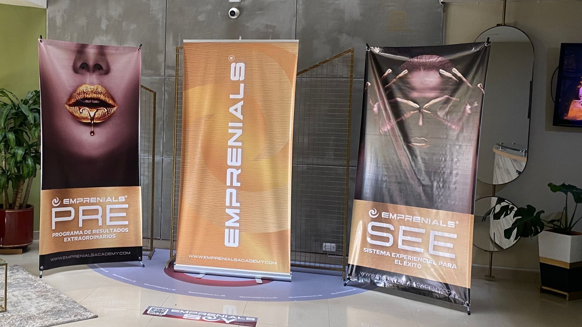 Three vertical banners with text and logos, placed in a lobby area with a tiled floor, a potted plant, and a mirror on the wall.