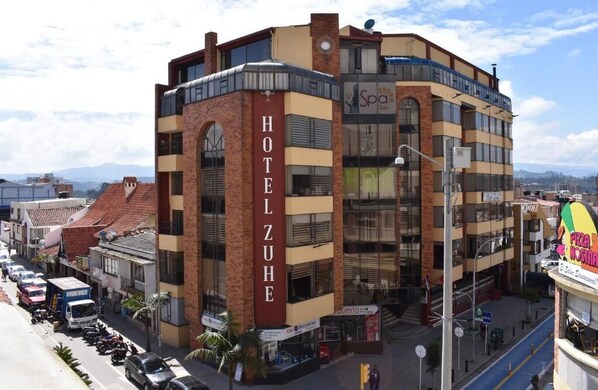 A multi-story hotel with a prominent 'HOTEL ZUHE' sign, featuring a modern design with brick and glass elements, located in an urban setting with visible streets and other buildings. - Hotel Zuhe (Paipa)