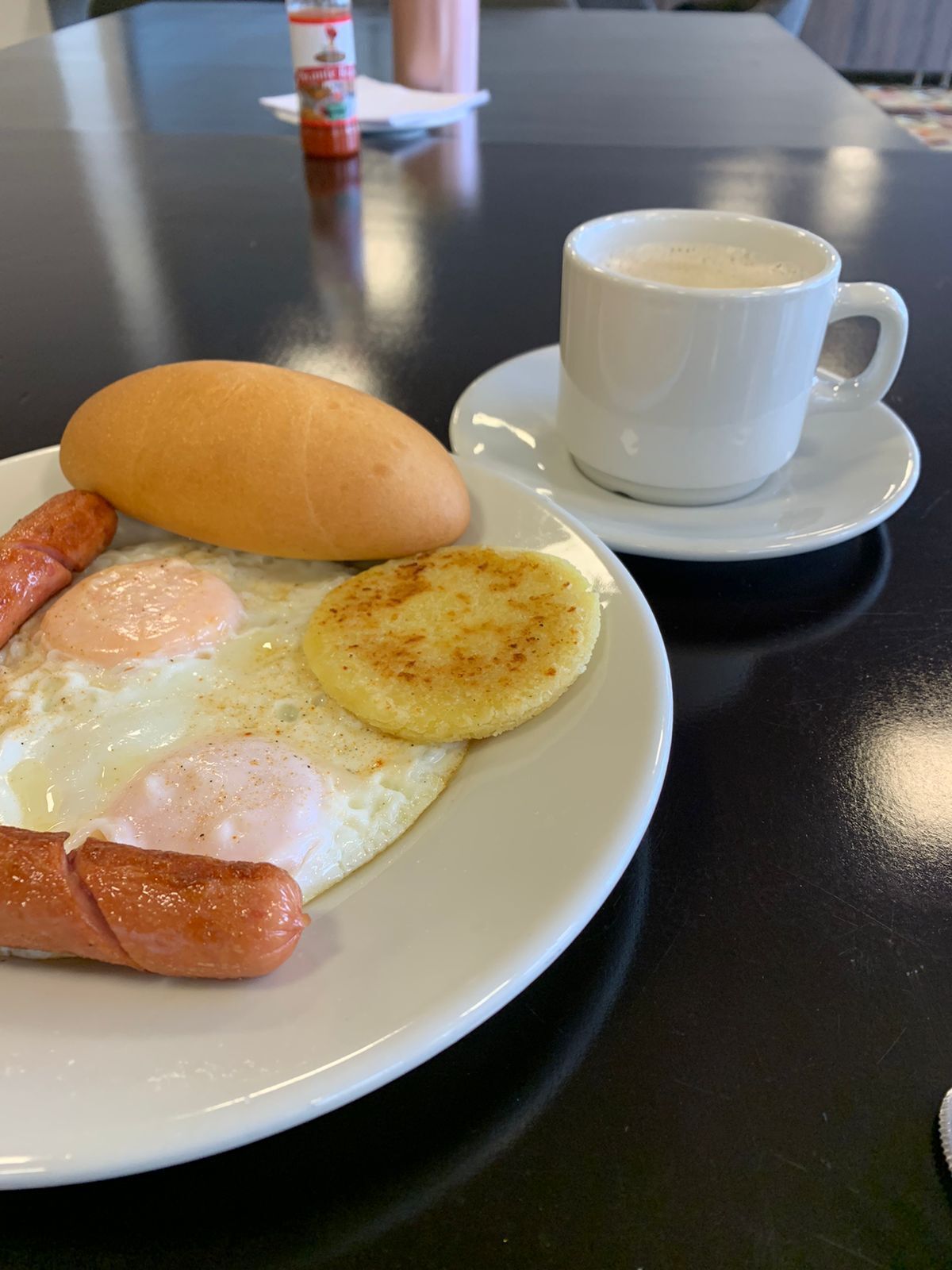 A plate with breakfast items including sausages, eggs, and a bread roll, accompanied by a cup of coffee on a table.