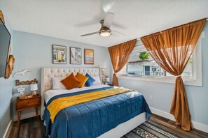 A bedroom with a tufted headboard, a bed with blue and yellow bedding, a ceiling fan, and brown curtains.