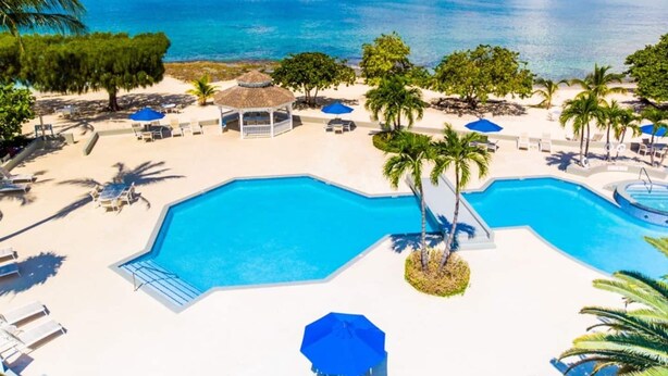 A resort pool area with clear blue water, surrounded by white sand, palm trees, and lounge chairs.