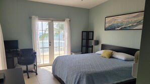 A bedroom with a bed, a desk, a chair, a TV, and a view of the beach through the window.