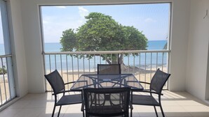 A balcony with a table and chairs, a view of the beach, and a tree.