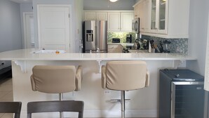 A modern kitchen with white cabinets, a stainless steel refrigerator, a built-in oven, and a bar area with two stools.